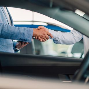 Zwei Personen geben sich in einem Autohaus die Hand, was einen erfolgreichen Geschäftsabschluss oder eine Vereinbarung symbolisiert. Im Vordergrund ist ein Teil des Fahrzeuginnenraums zu sehen.