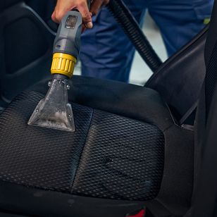 Eine Person, die einen Staubsaugeraufsatz verwendet, um einen strukturierten schwarzen Autositz gründlich zu reinigen, wobei sie sich darauf konzentriert, Schmutz und Ablagerungen zu entfernen.