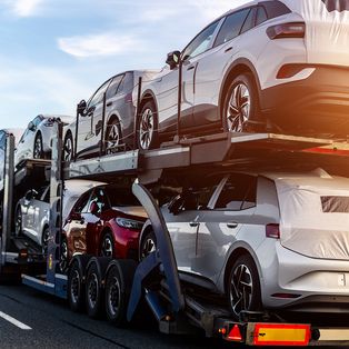 Ein Autotransporter mit mehreren neuen Elektrofahrzeugen, darunter ein weißer SUV oben und ein rotes Auto unten, vor einem klaren Himmel.