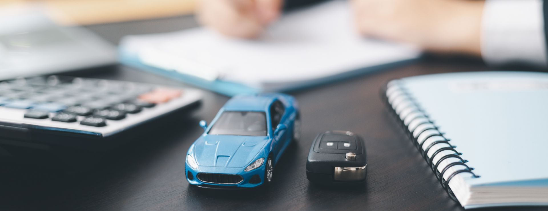 Ein blaues Spielzeugauto und ein Autoschlüssel liegen auf einem Schreibtisch neben einem Taschenrechner und einem Notizbuch, während im Hintergrund eine Person schreibt.